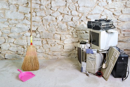 Inside a room or garage space with a textured stone wall in the background, there is a collection of discarded electronic waste, including an old computer monitor with a beige frame, a small white and grey desktop tower, and a keyboard lying on the ground. Several cables are tangled among the electronic devices. To the left, a broom with a wooden handle and straw bristles leans against the stone wall, and a bright pink dustpan made of plastic is placed on the floor nearby. The floor appears to be plain concrete, and the scene suggests a storage or disposal area for rubbish, relevant to rubbish removal services such as those offered by Business Waste Removal Crystal Palace. The arrangement indicates an effort to clear away old appliances and cleaning tools, with the electronic waste possibly awaiting collection or disposal, aligning with local waste management needs in the Crystal Palace area.