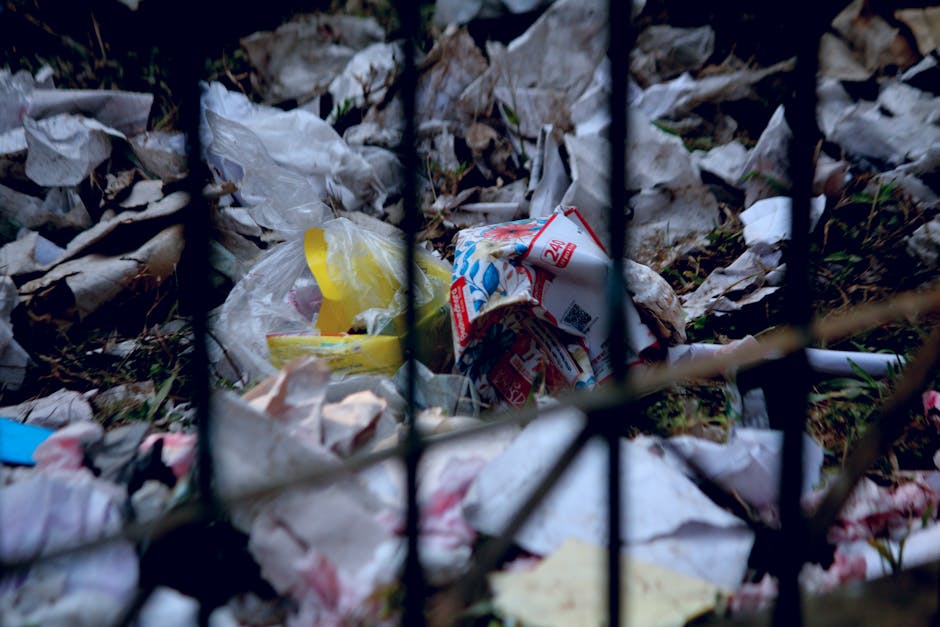 A close-up image of discarded aluminum cans and plastic beverage containers lying on a paved surface. The cans are predominantly gold-colored with silver tops and pull tab openings, some showing resid
